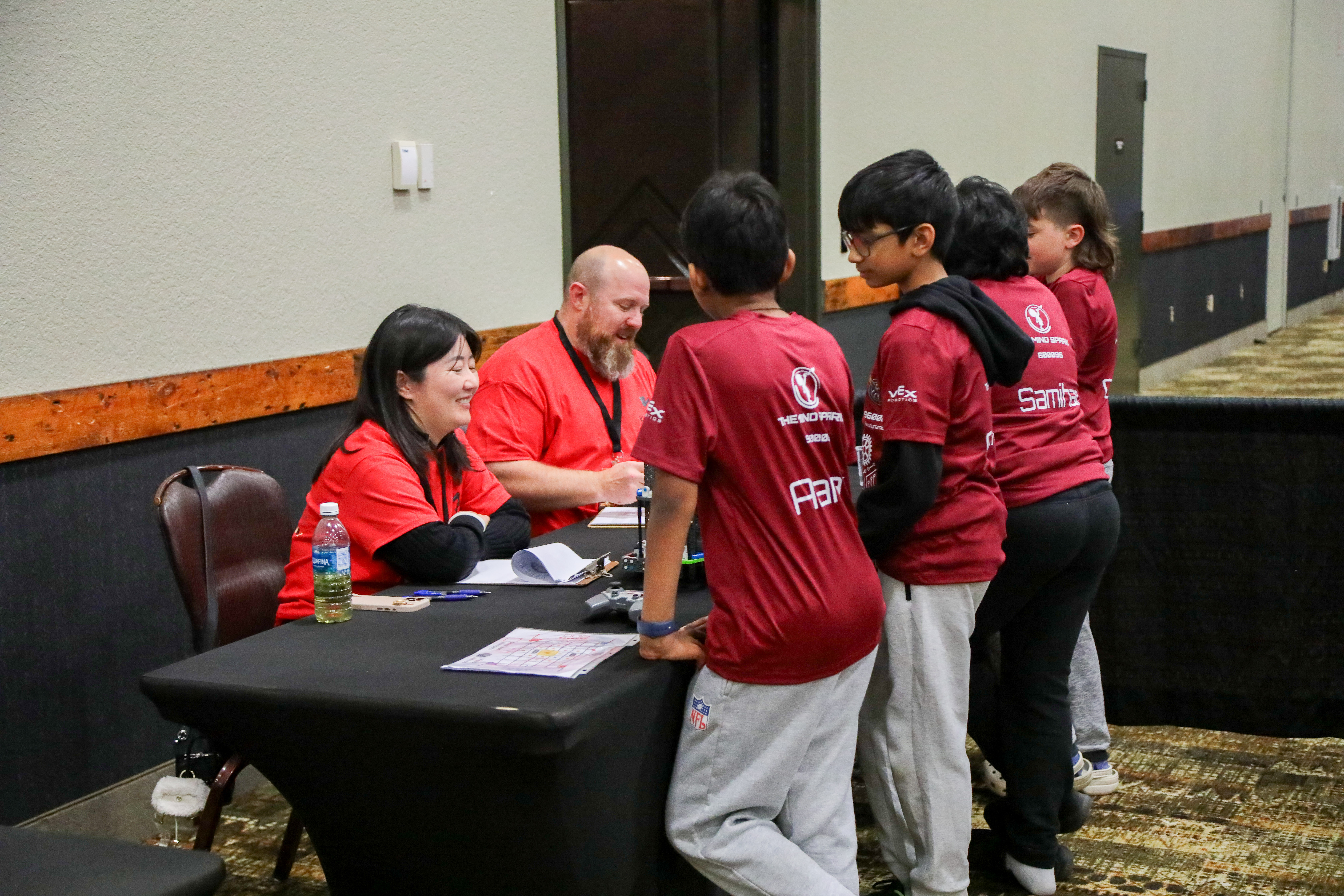 A pair of judges speaks with a team at a table, talking about their robot and taking down notes on their clipboards.