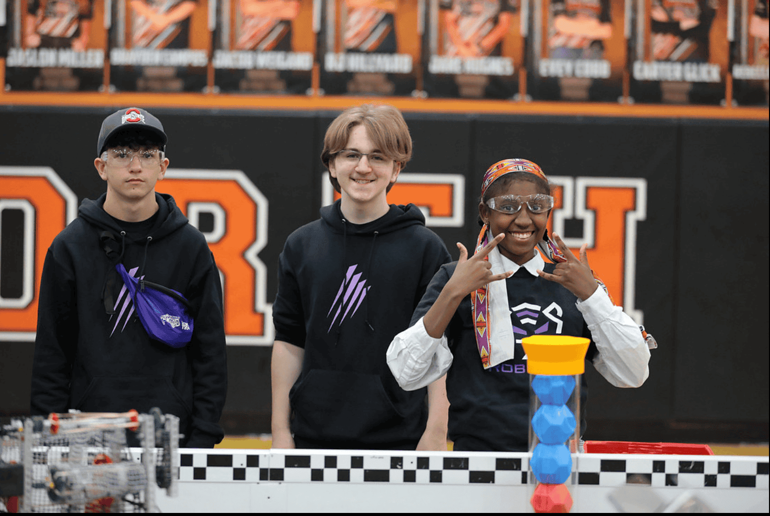 Three members of a V5 team stand beside a Push Back field at a competition. One is smiling and making the I love you symbols with their hands