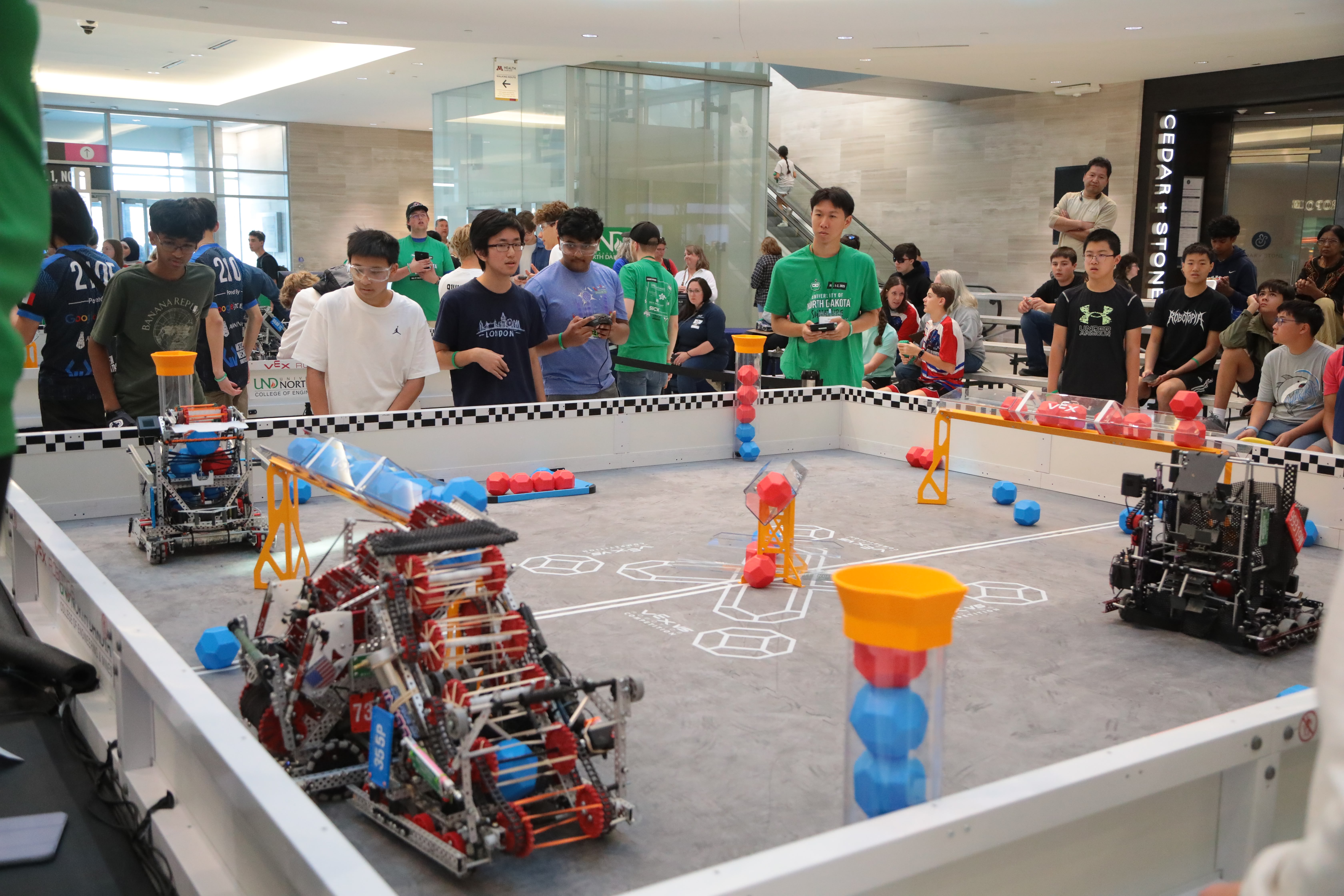 Team members, judges and refs are assembled around a V5 Competition field with three robots. One boy holds a V5 controller in his hand.