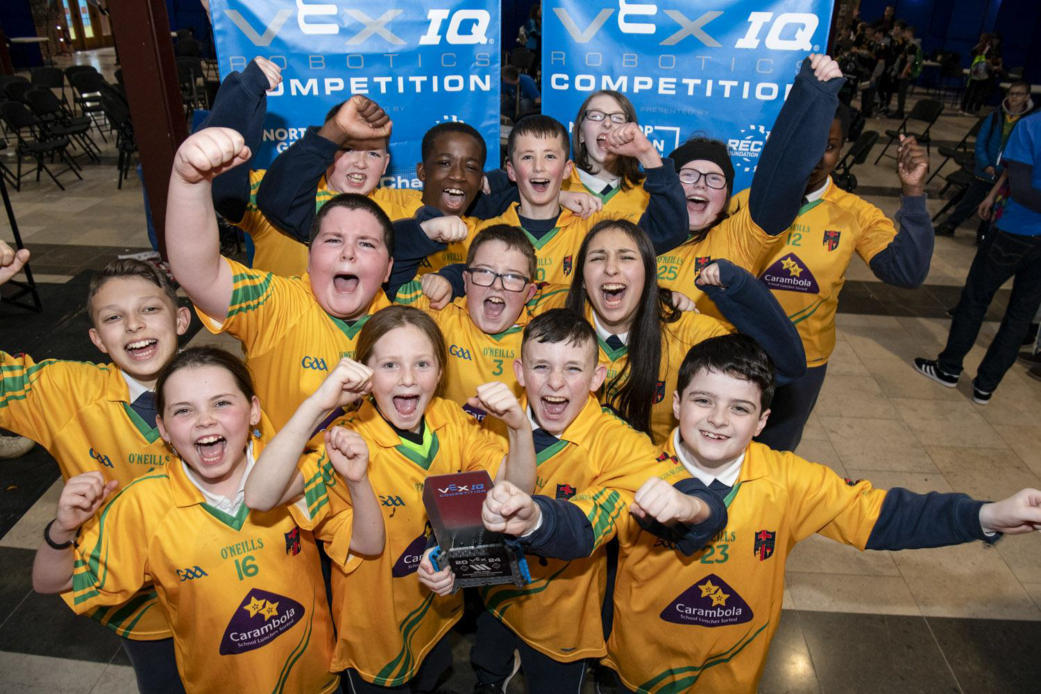 A team of students cheers and celebrates with their award in front of VEX IQ Robotics Competition banners.