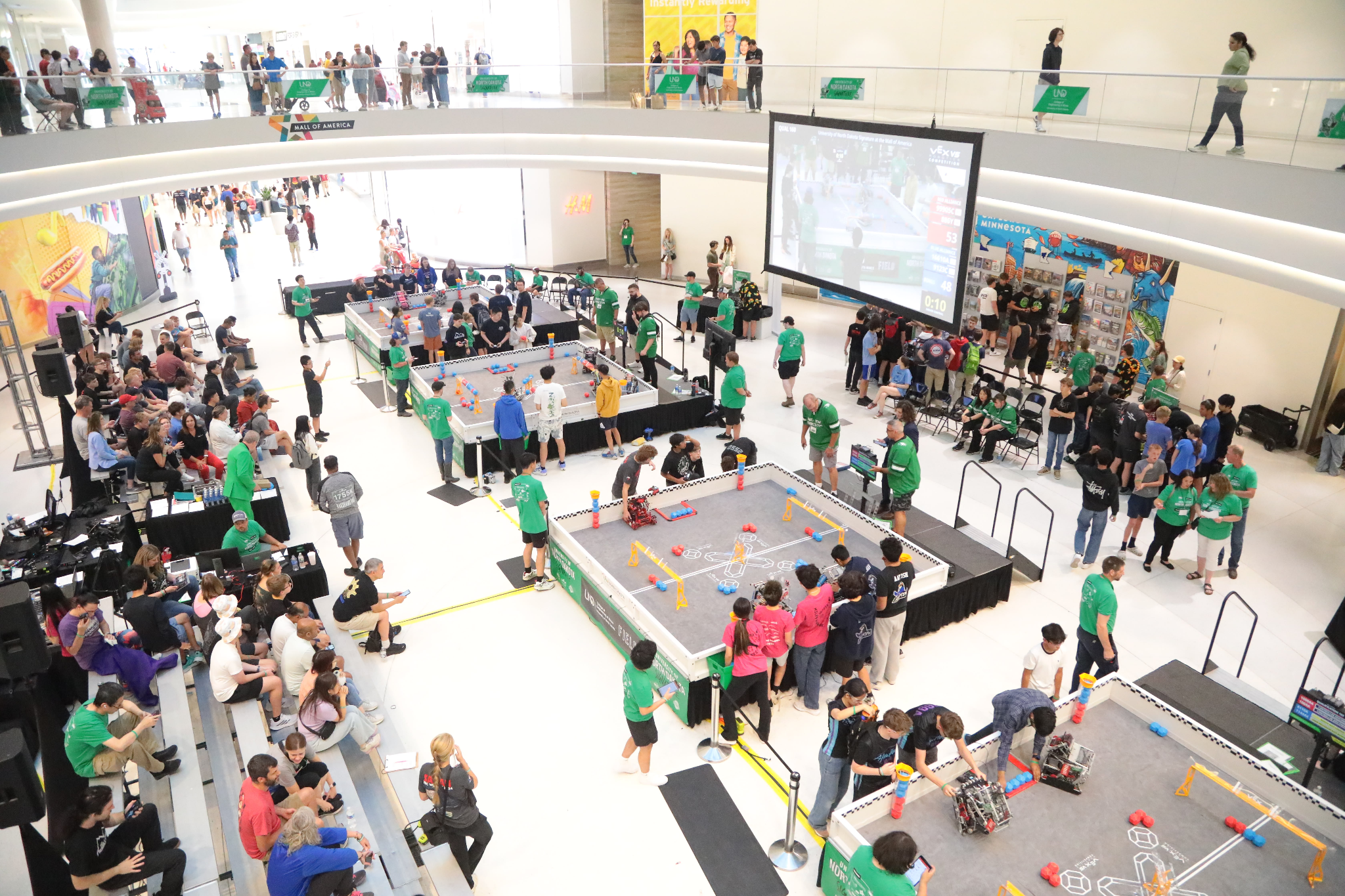 Overhead view of a VEX Robotics Competition event in progress, with multiple competition fields surrounded by teams, volunteers, and spectators.