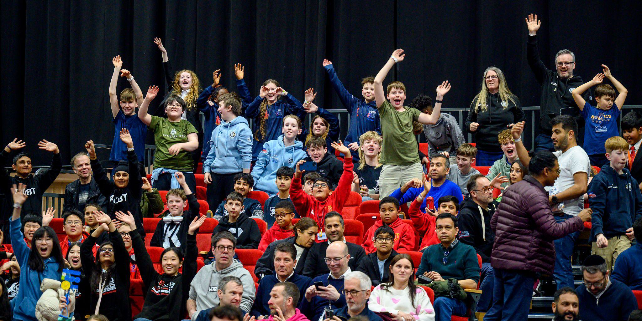 Students and mentors cheer from arena seating at a VEX Robotics competition.