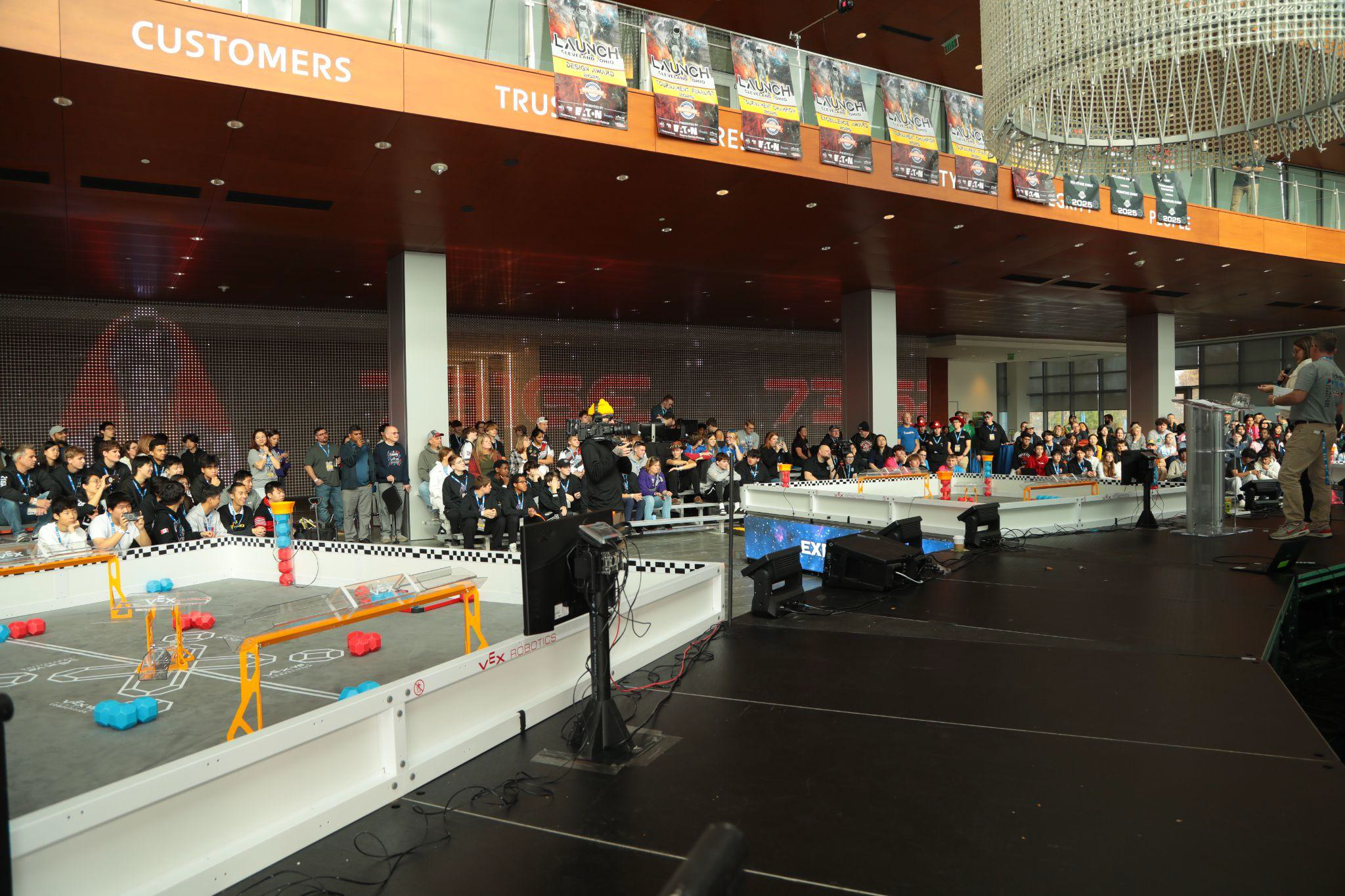 Spectators watching a match on VEX Robotics Competition fields set up in a large event venue.