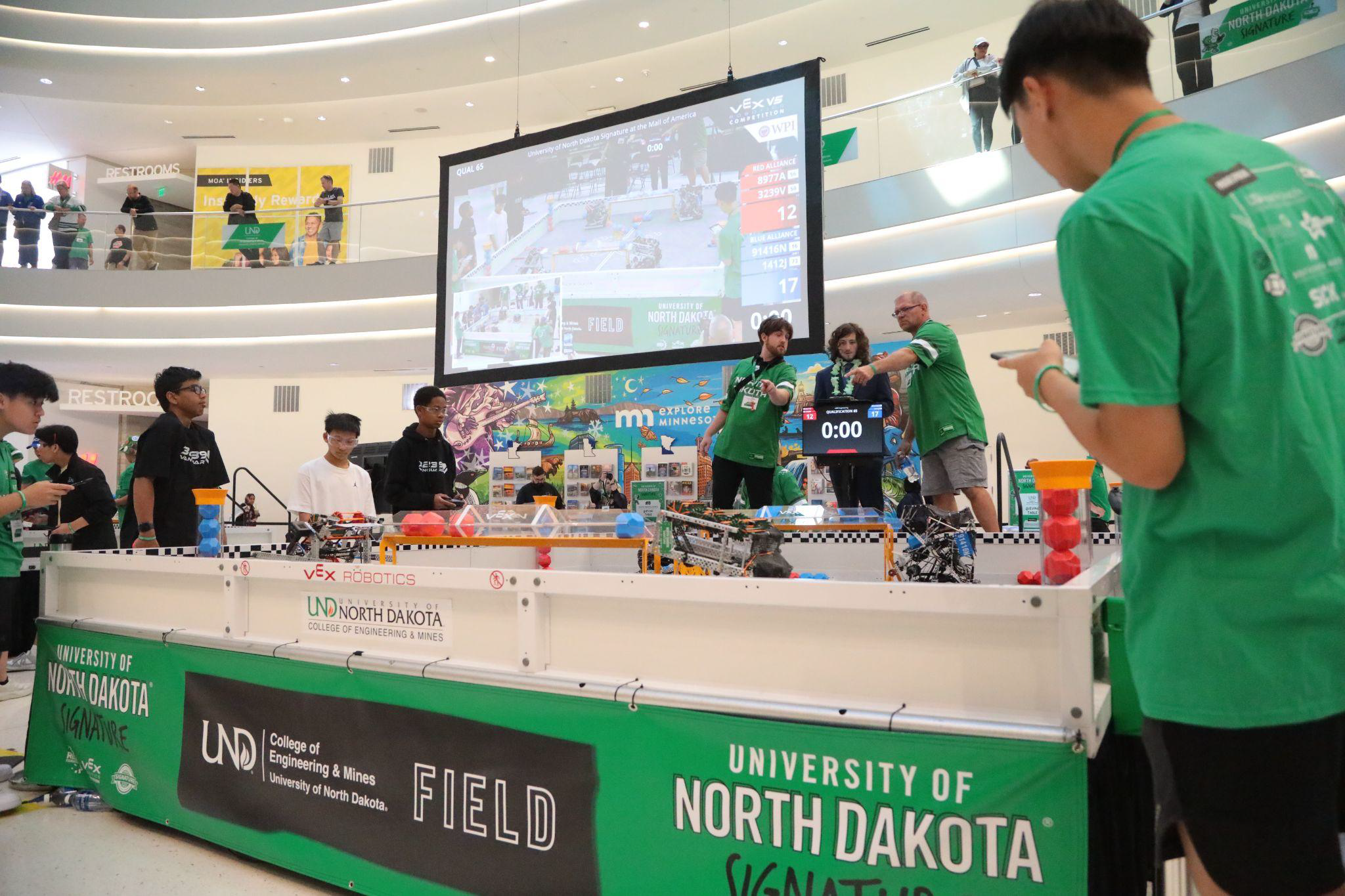 Students beside a VEX Robotics Competition field at a large event, with an overhead screen showing the live match.