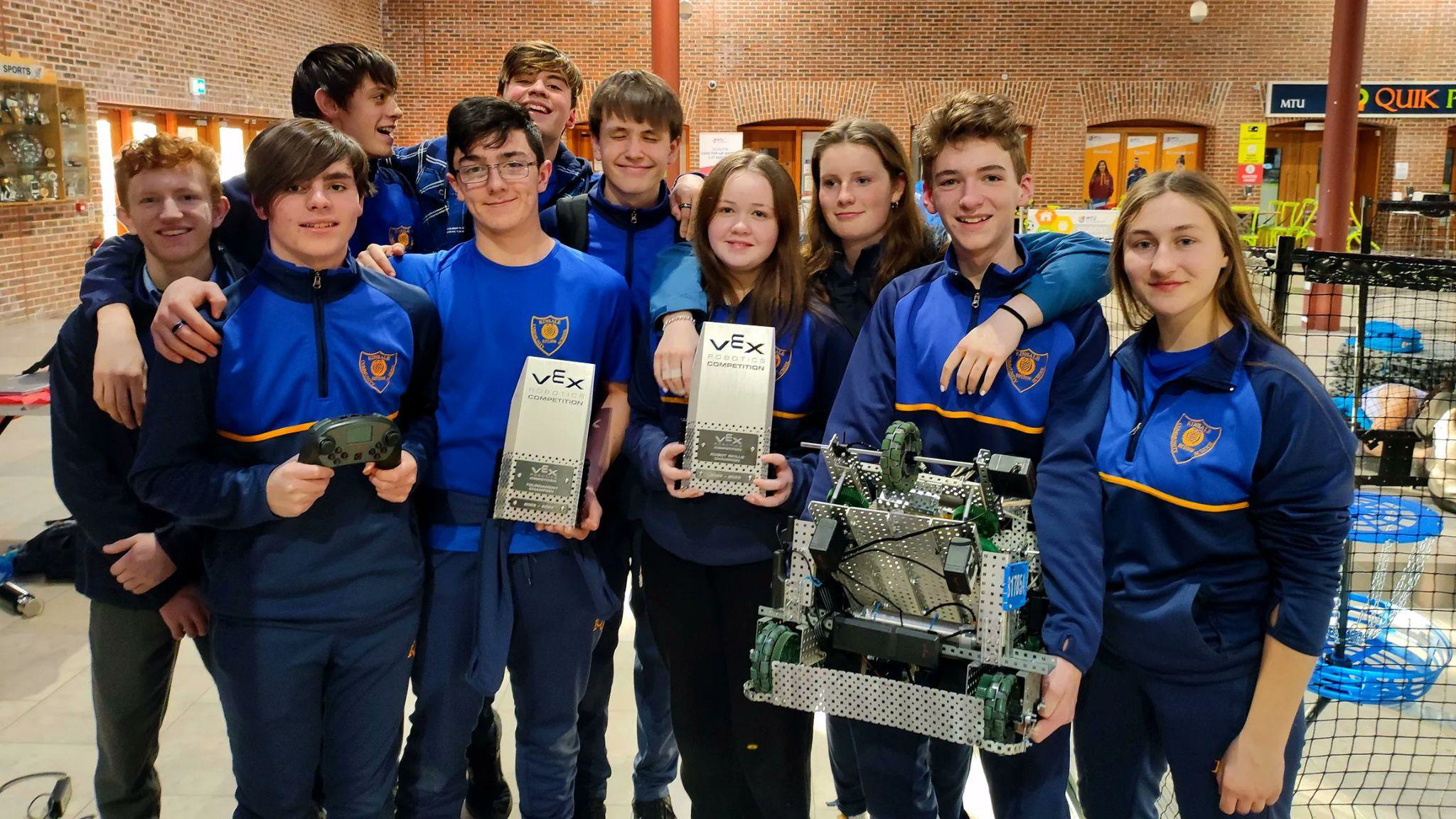 A team in blue school uniforms poses with their robot and awards after a competition.
