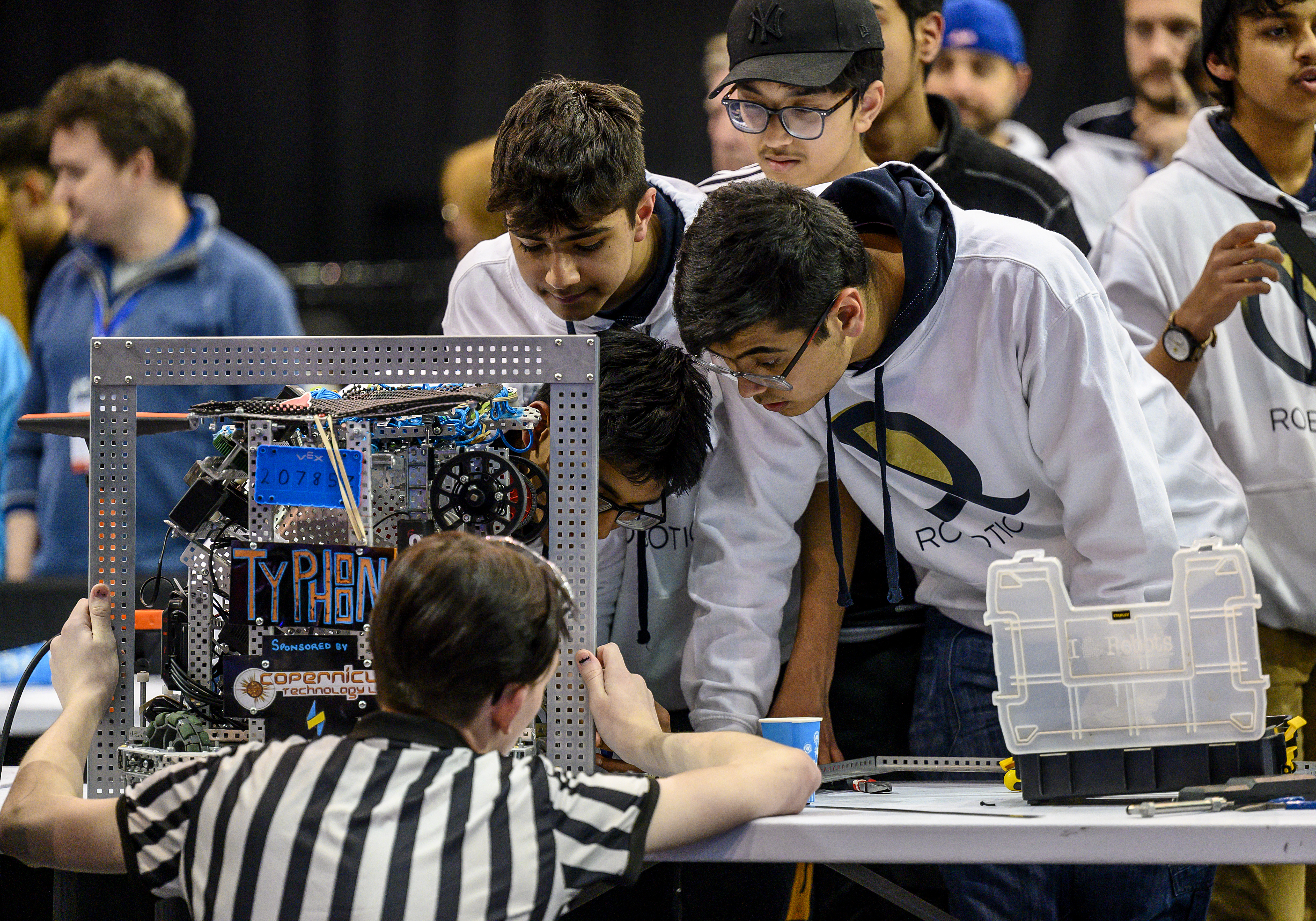 One group of students standing on one side of the table looking down at a robot on the table. On the other side of the table, an inspector crouches while holding a metal sizing tool around the robot. They all are looking to see if the size of the robot fits within the metal sizing tool.