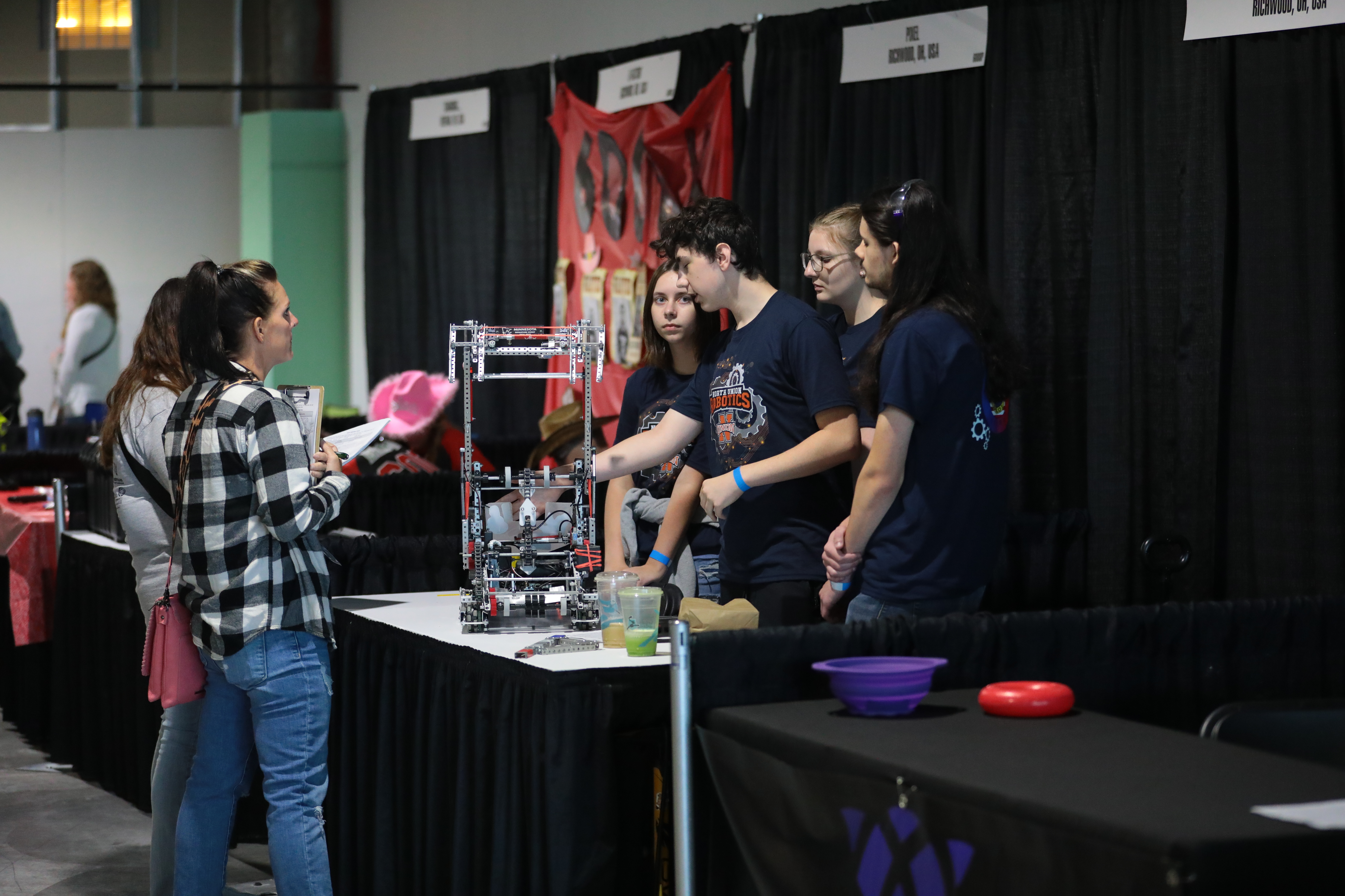 A team stands at their pit talking to two judges with clipboards. One student on the team points to a feature of the robot while talking ,and the other girls look on listening.