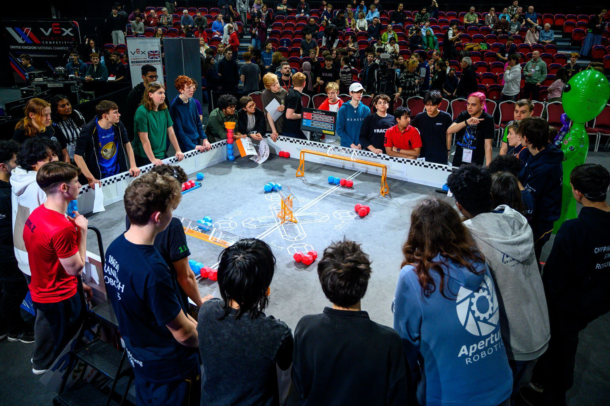 Students gathered around a VEX Robotics competition field watching a match at a tournament.