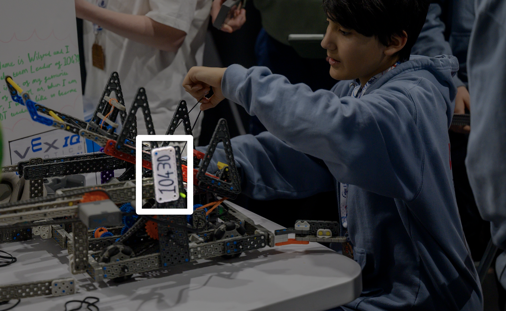 An IQ robot sits on a table in a pit as a student works on it. The white license plate on its side with the team number is highlighted.