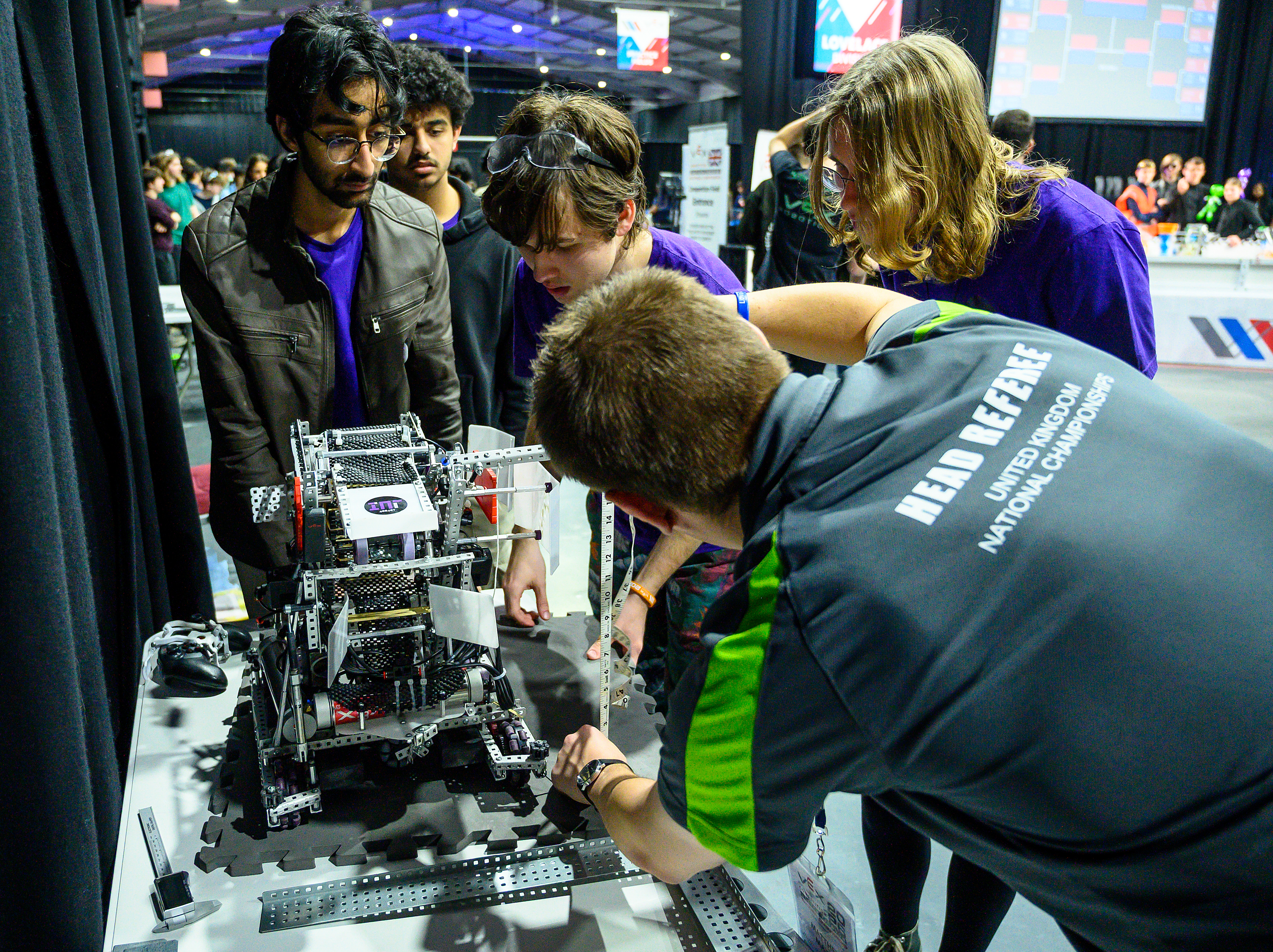 One group of students standing on one side of the table looking down at a robot on the table. On the other side of the table, an inspector crouches while holding a tape measure to inspect the sizing of the robot.