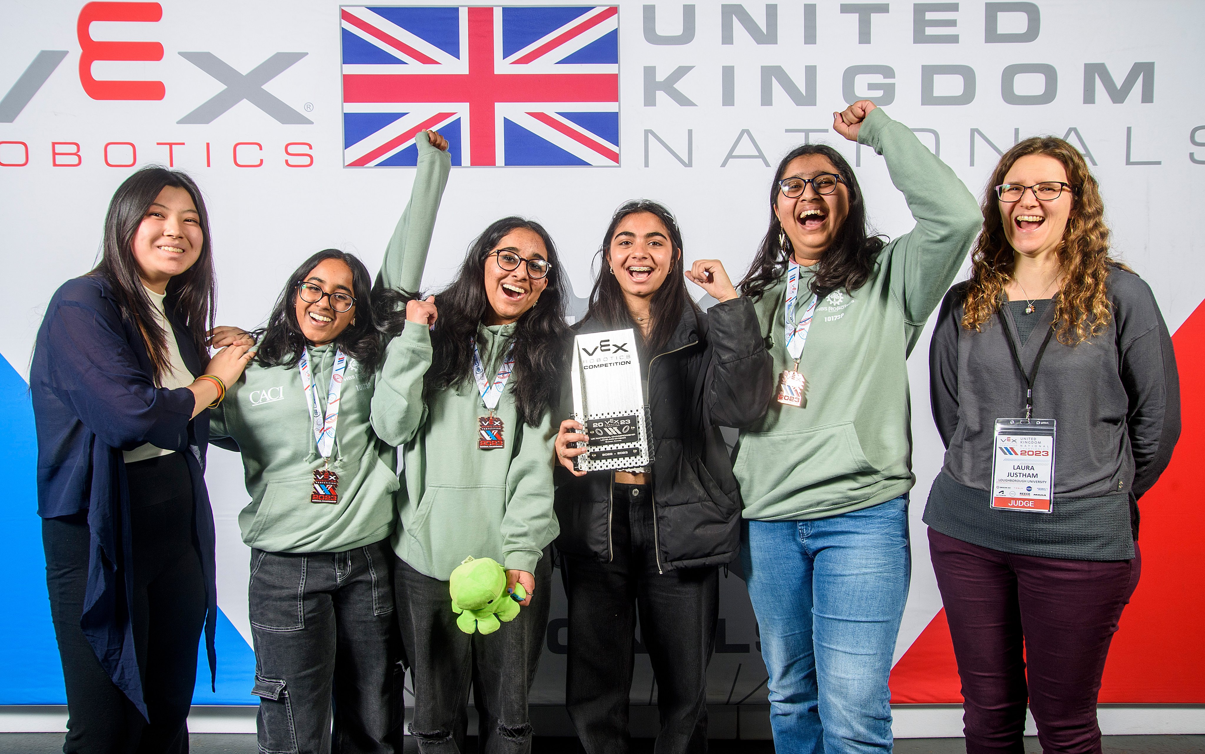 A team cheers in front of a competition banner, holding their award. A smiling judge stands alongside the team.