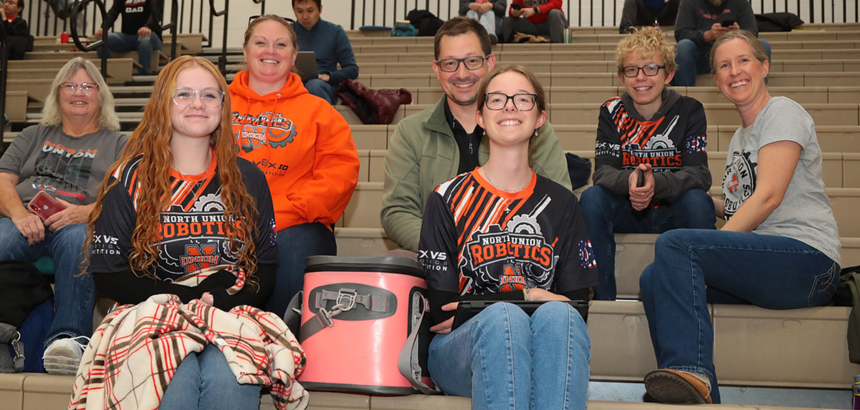 A group of parents for a VEX Robotics team sitting on the bleachers at a VEX Robotics Competition.
