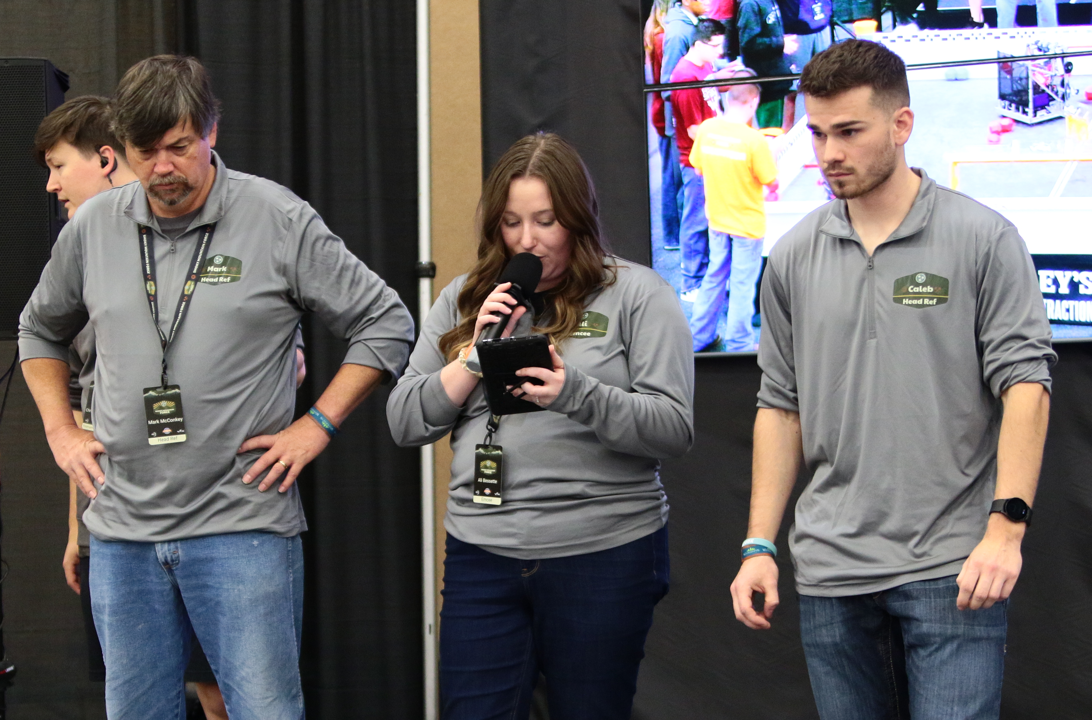Three adults watching a VEX Robotics competition, with a screen showing the competition running behind them.