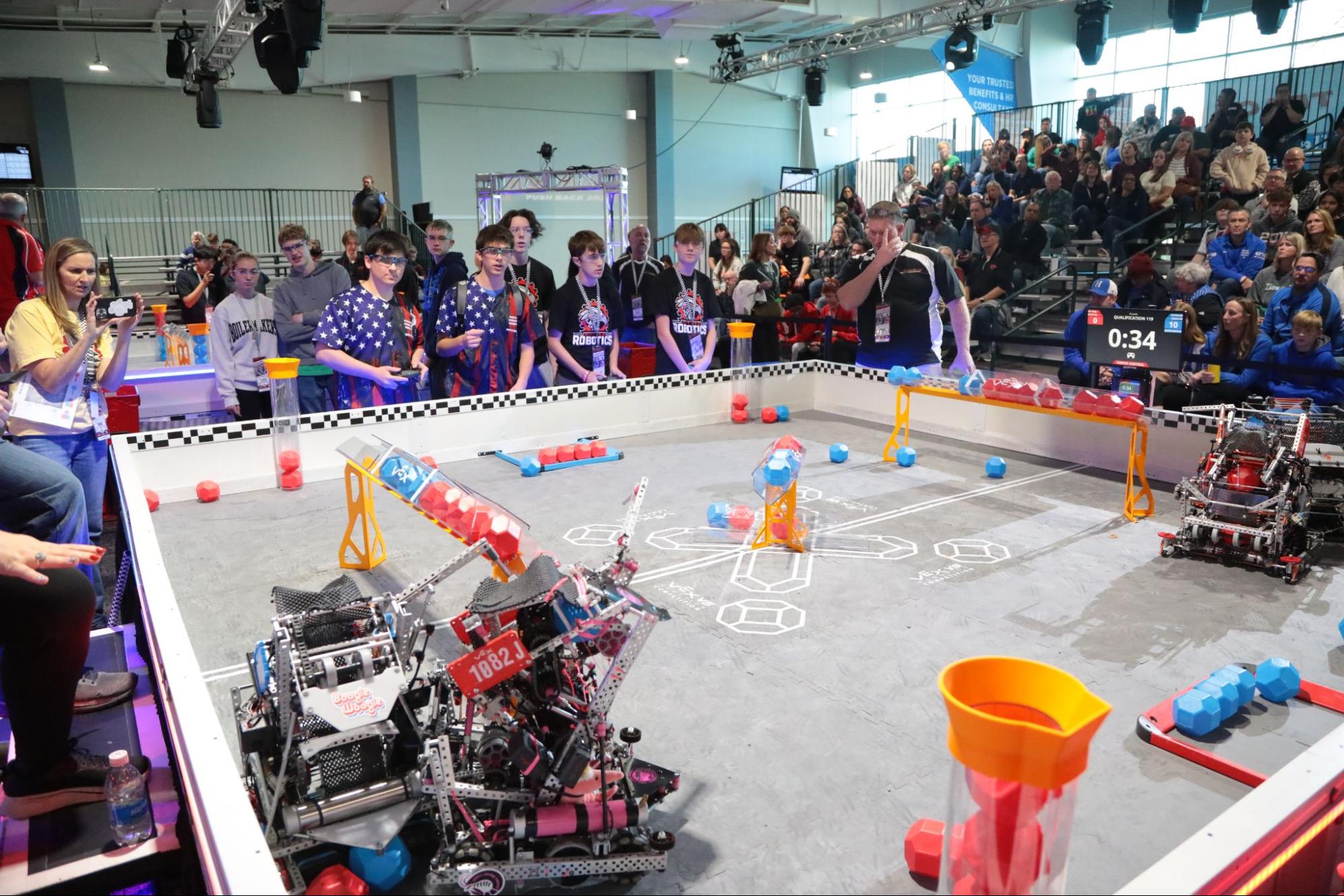 A competition field with a live V5 match happening. Behind the field in the foreground, spectators sit in bleachers. On the side, a coach videos the match on a phone. A referee is counting beside the drive team. 